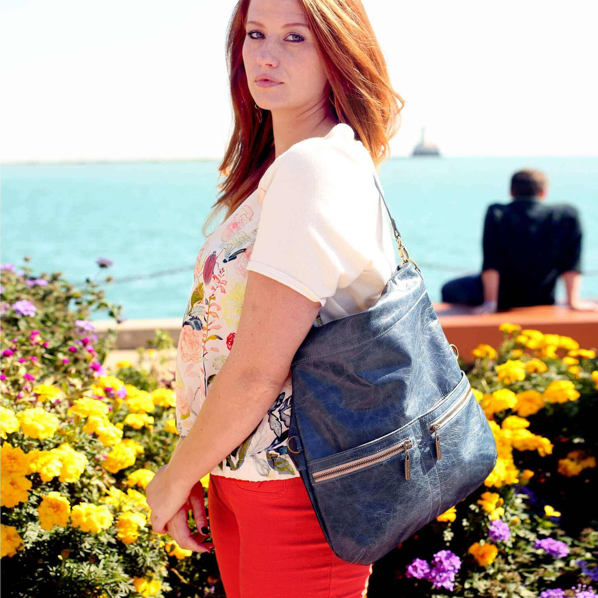 Model showcasing Mini Lauren 6-in-1 convertible leather crossbody bag by vibrant flowers near water.