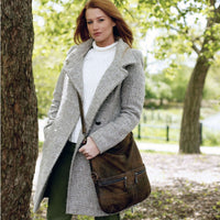 Woman in a coat holding a leather crossbody bag outdoors surrounded by greenery.