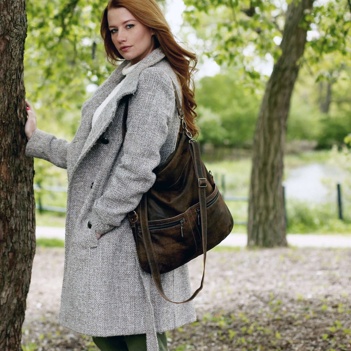 Woman wearing a grey coat with the Original Lauren leather crossbody bag outdoors, surrounded by greenery.