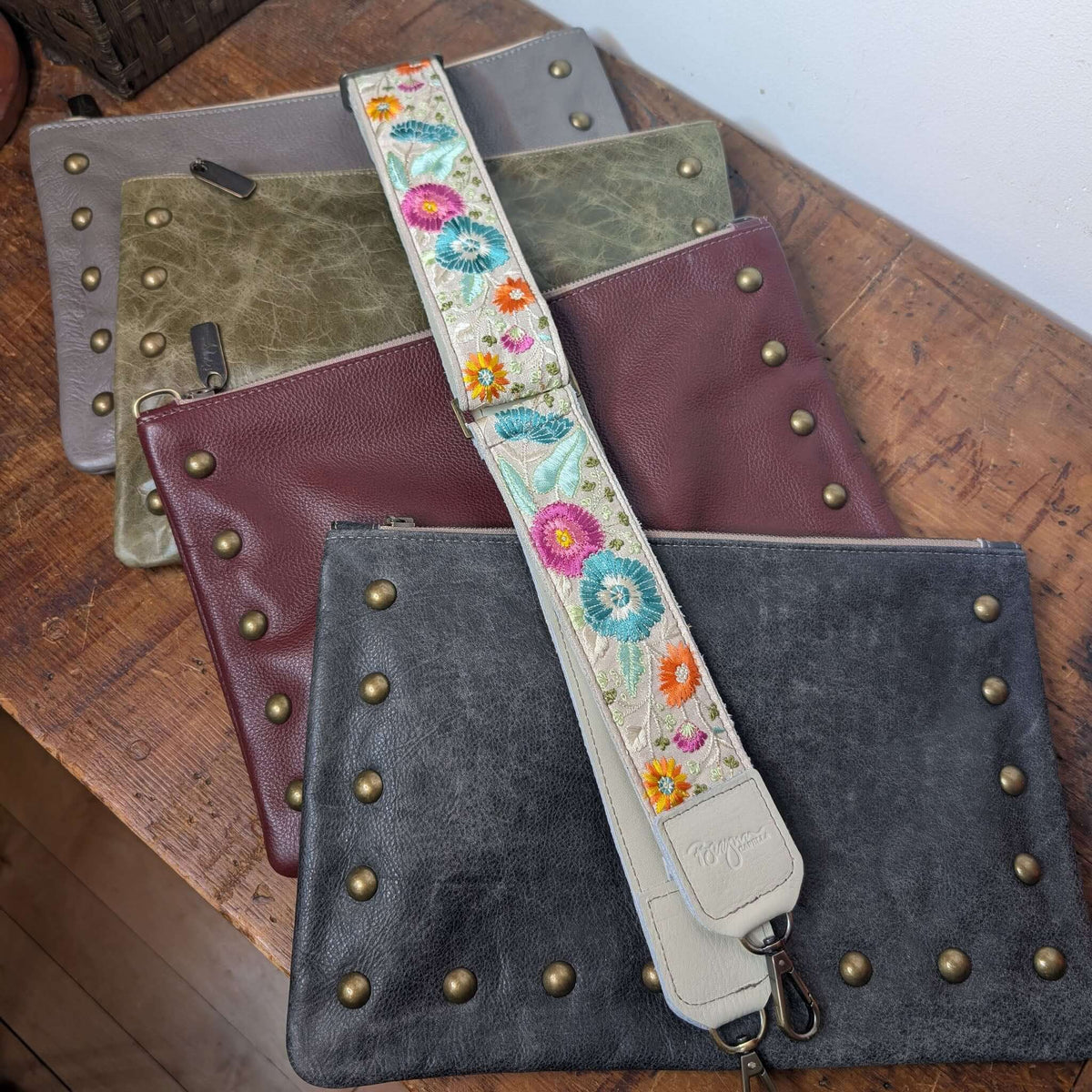 Lizzy Guitar Bag Strap - Laguna Wildflowers shown with various handbag clutches on a wooden surface.
