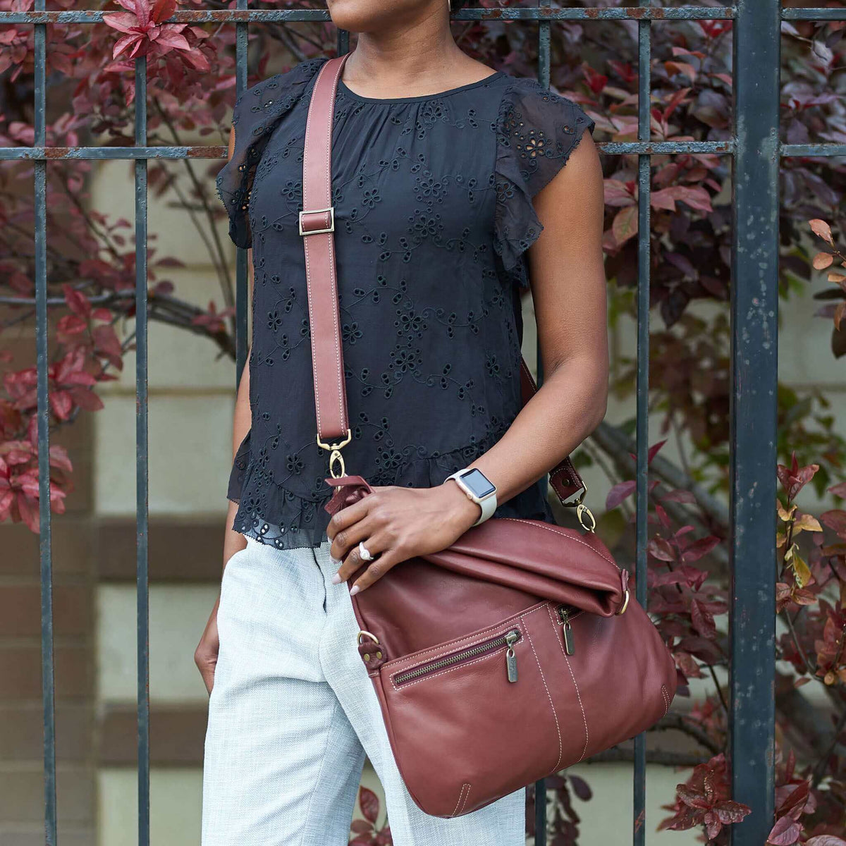 A woman wearing a black top and gray pants holds the Original Lauren leather crossbody bag in Merlot.