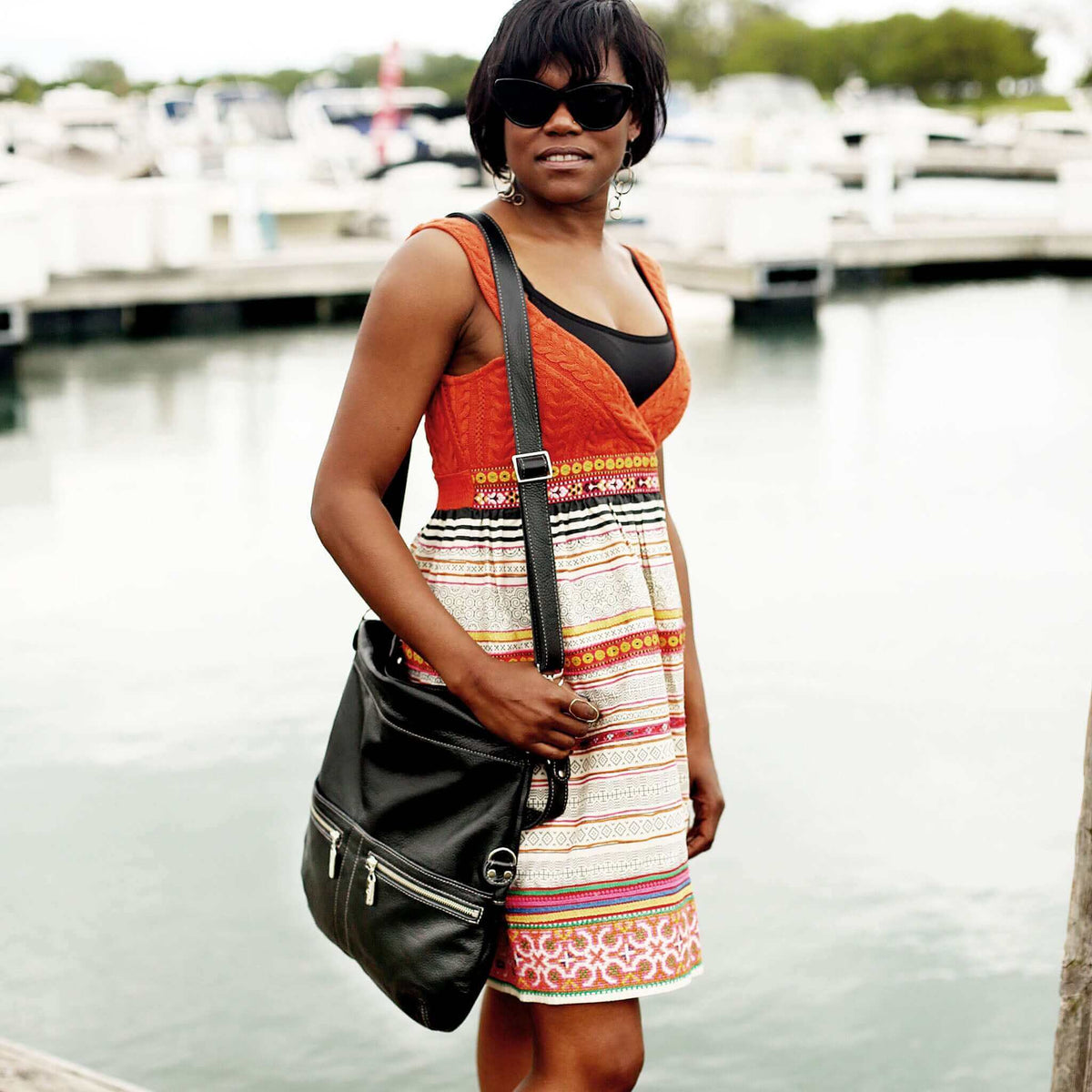 Woman wearing a colorful summer dress and sunglasses, carrying a leather crossbody bag by the water.