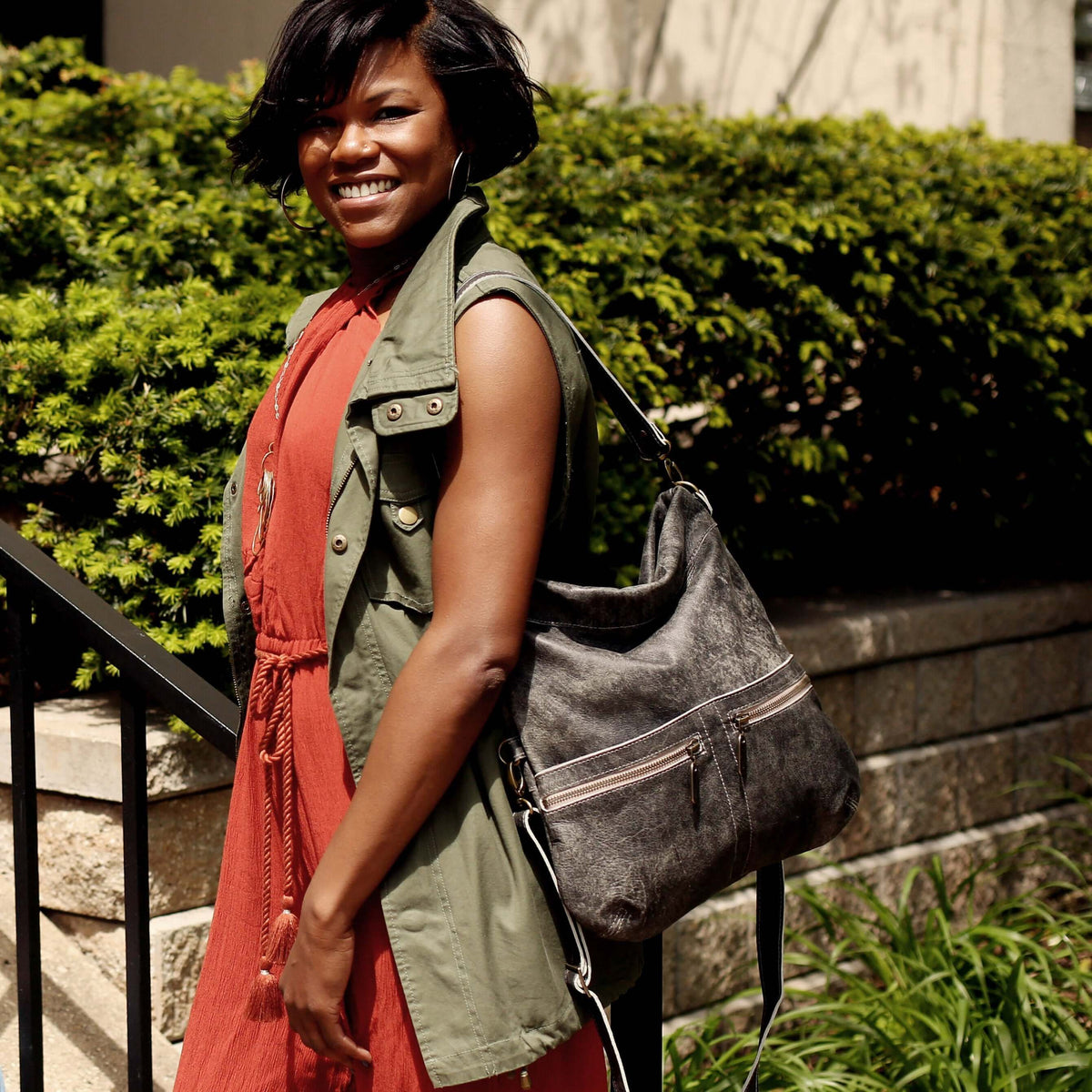 Woman wearing a green vest and red dress, showcasing a leather crossbody bag while posing outdoors.